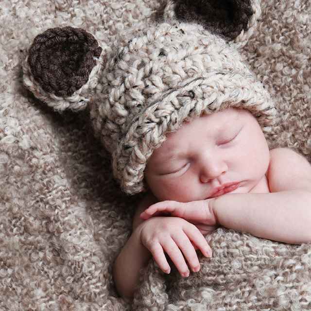 Photo of a newborn baby wearing teddy bear ears as photo accessory, nestled in a cozy blanket.