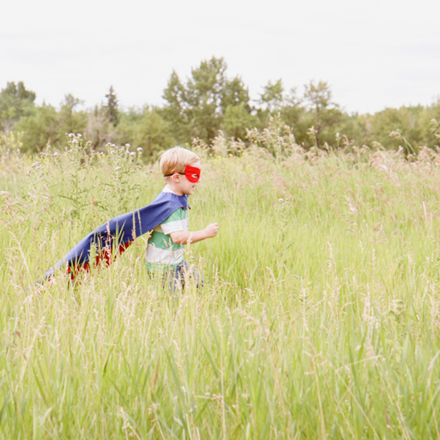 Photo of a child running in a wheat field with his superman cap.