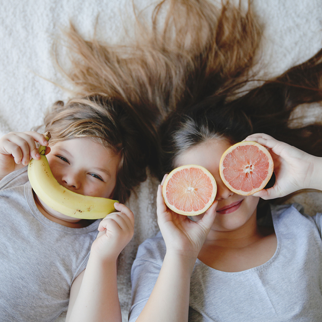 Photo of two girls in bed playing with fruits.