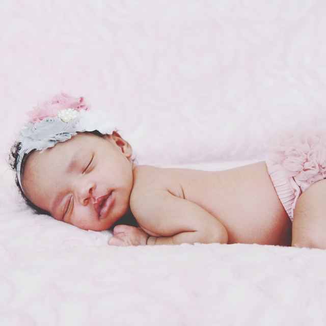 Photo of a newborn sleeping in bed and wearing a fabric flowers headband.